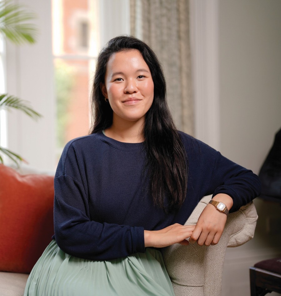 Headshot of Jackie Yip, a woman wearing a long sleeve navy sweater and a teal skirt, sitting on a tan couch.