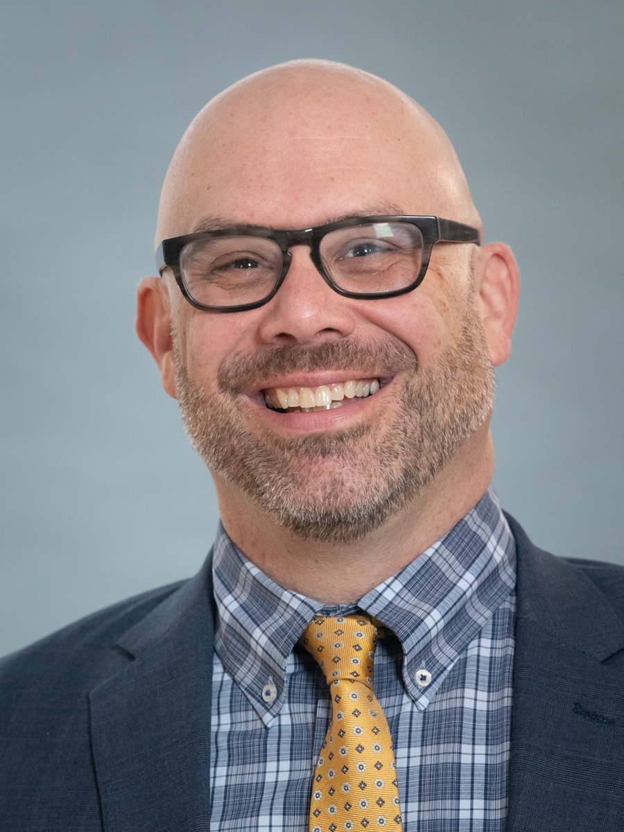 Headshot of Keith Paul, who wears glasses, a blue checked button down, yellow tie, and dark blue blazer.