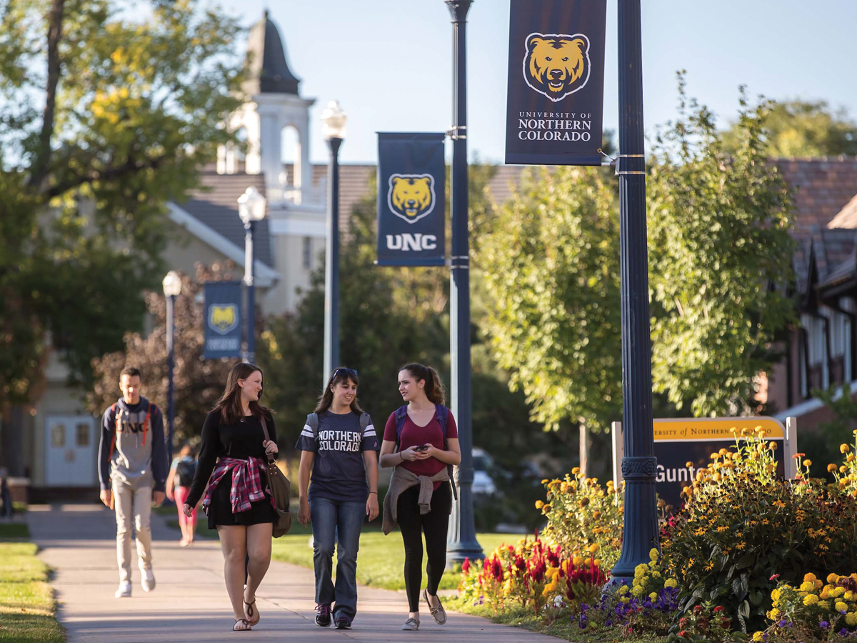 On the University of Northern Colorado campus, three students walk together down a path. The middle student wears a UNC-branded shirt.