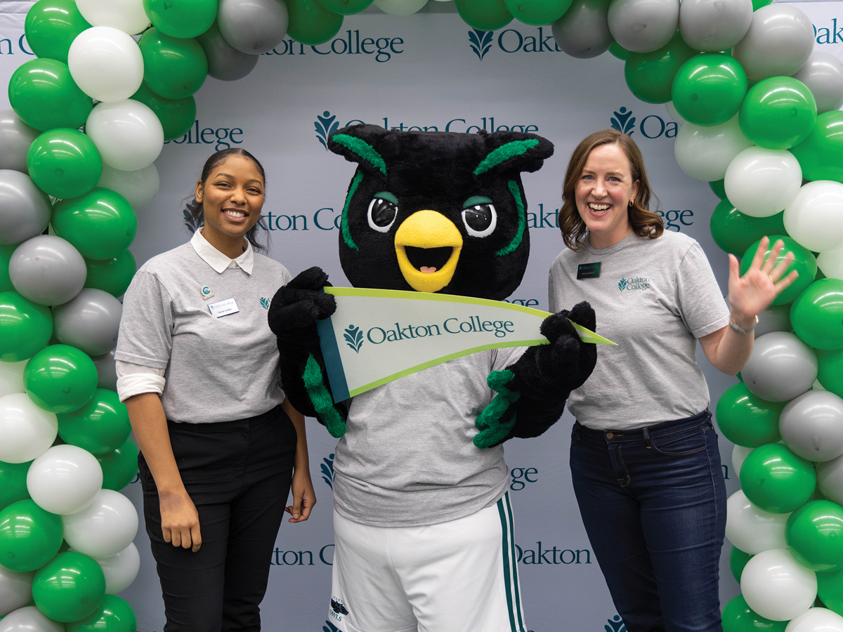 Two women pose on either side of a green bird mascot, a green/gray/white balloon arch around them. They all wear Oakton-branded shirts and the mascot holds an Oakton-branded pennant.