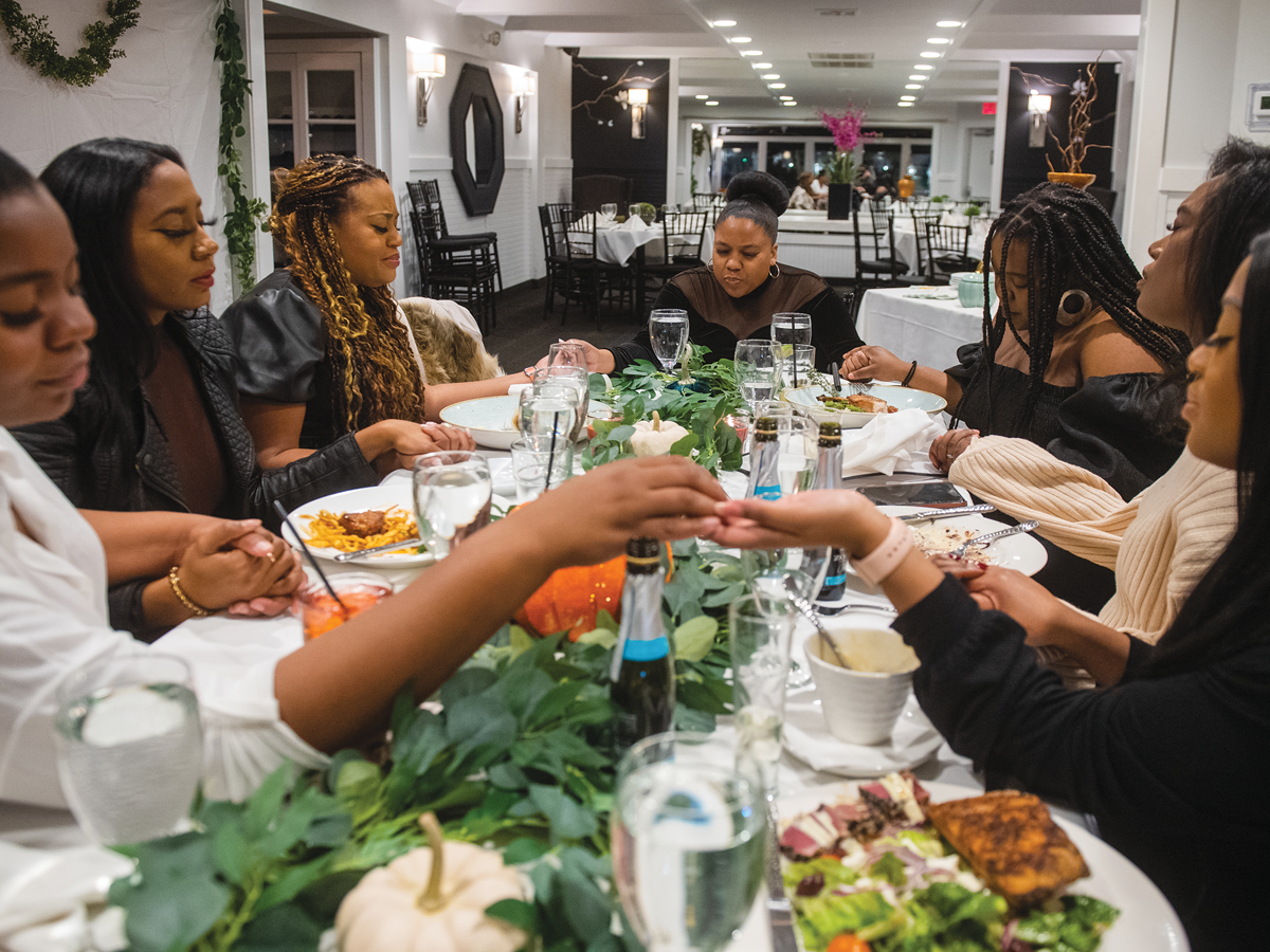 A group of Black women hold hands around a white tablecloth covered table full of food, eyes closed in prayer.