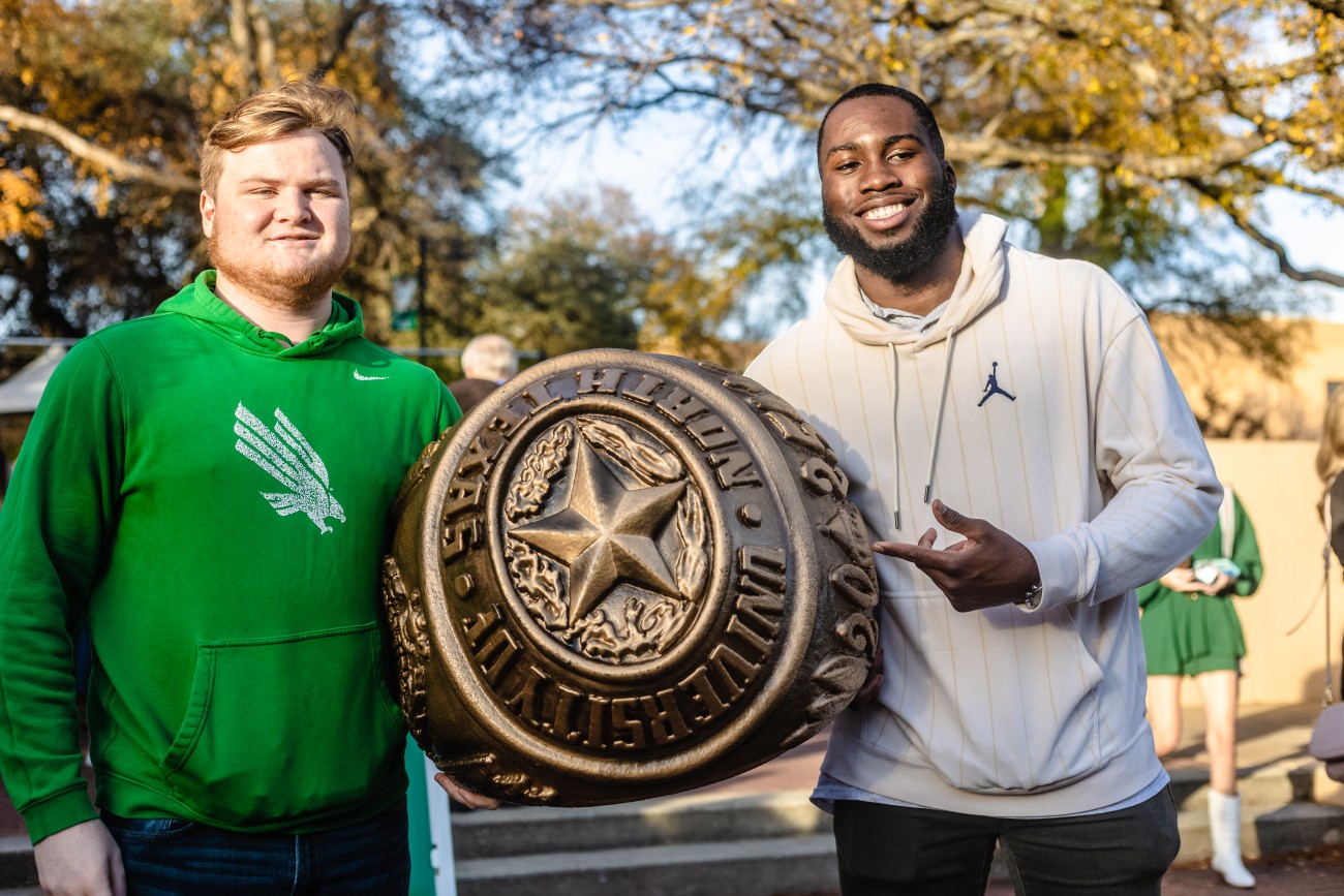 Two University of North Texas graduates stand on either side of a bronze statue of a UNT class ring.