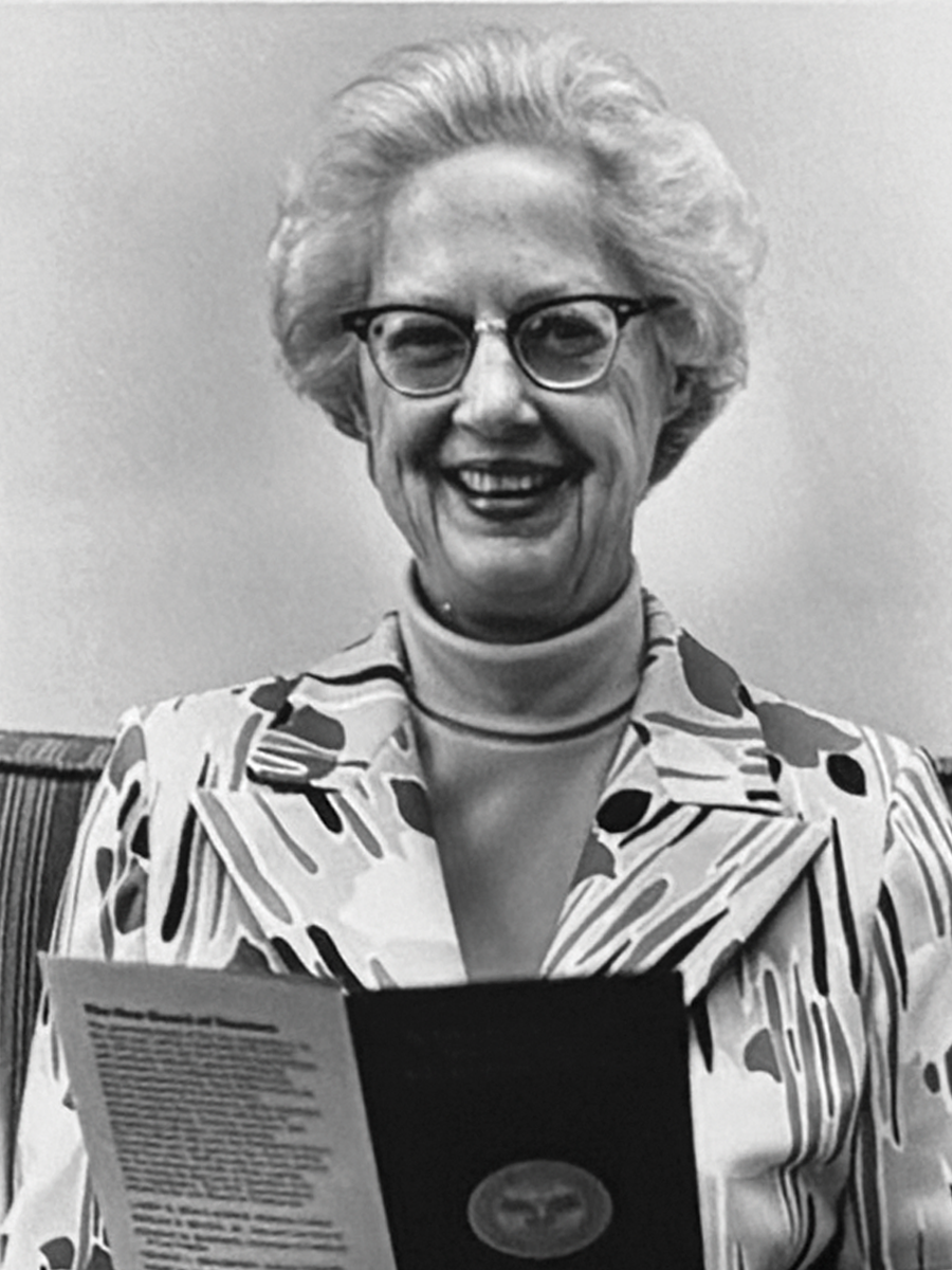 Photo of Alice Beeman. She wears a turtleneck and patterned blazer and holds a pamphlet open in front of her.